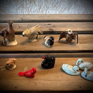 May include: A collection of nine ceramic bird figurines, including a cardinal, a swan, a black bird, a parrot, a turkey, a dove, and an eagle. The figurines are painted in various colors, including red, white, black, brown, and blue.