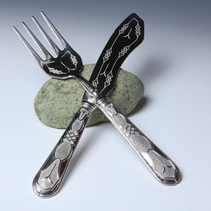 May include: Silver fork and knife set with ornate, engraved designs. The fork and knife are crossed over a small, green stone. The handles and the blade of the knife feature detailed patterns, adding to the elegance of the cutlery.