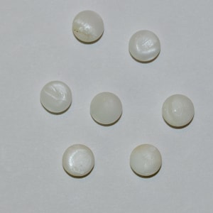 May include: Seven small, round, off-white beads are arranged on a white surface. The beads have a slightly iridescent sheen, suggesting a pearl-like quality. The arrangement is scattered, with each bead distinct.