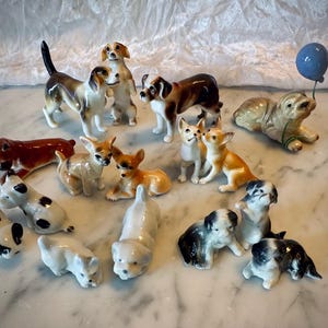 Könnte beinhalten: Eine Sammlung von 15 Porzellan-Hundefiguren in verschiedenen Posen und Rassen, darunter ein Chihuahua, ein Mops und ein Dackel. Die Figuren sind in realistischen Farben bemalt und haben komplizierte Details. Einige der Figuren halten einen blauen Ballon.
