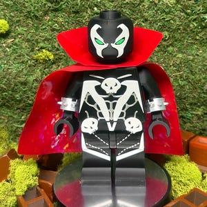 Puede incluir: Figura de acción en blanco y negro con una capa roja. La figura tiene un diseño de calavera en el pecho y una cara blanca con ojos verdes. La figura está sobre una base plateada.