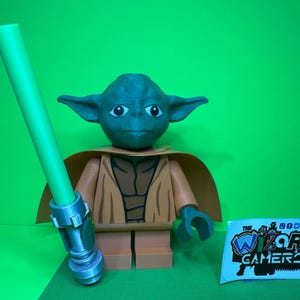 May include: A green lightsaber and a green and brown Yoda Lego minifigure. The minifigure is wearing a brown robe and has a green face. The lightsaber is in the minifigure's left hand.