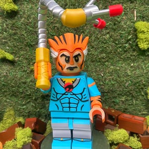 Puede incluir: Una figura Lego personalizada de un personaje de dibujos animados con una cabeza a rayas naranja y negra, torso azul y piernas grises. La figura sostiene un arma amarilla y plateada con puntas rojas. La figura está sobre una superficie reflectante.