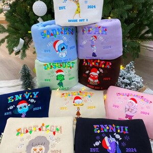 May include: A stack of embroidered sweatshirts featuring characters and words from the movie "Inside Out 2." Each sweatshirt is a different color, with designs including "Joy," "Sadness," "Fear," "Anger," "Disgust," "Envy," "Anxiety," "Embarrassment," "Nostalgia," and "Ennui."