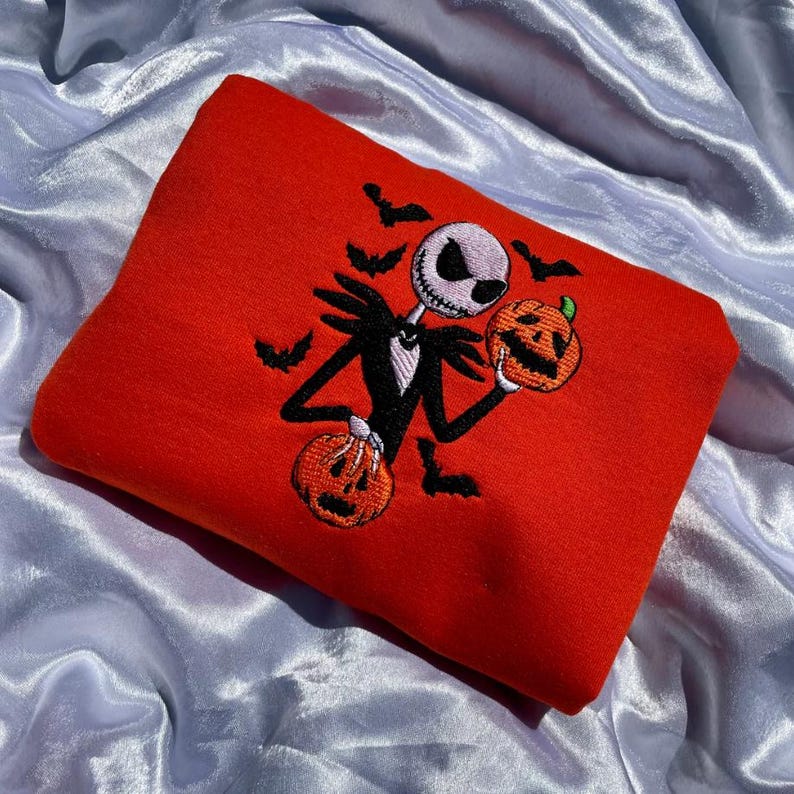 K&ouml;nnte beinhalten: Ein leuchtend rotes Sweatshirt mit einem gestickten Design von Jack Skellington, der zwei geschnitzte K&uuml;rbisse h&auml;lt. Schwarze Flederm&auml;use sind um die Figur herum gestickt. Das Design ist in Schwarz, Wei&szlig; und Orange gehalten.