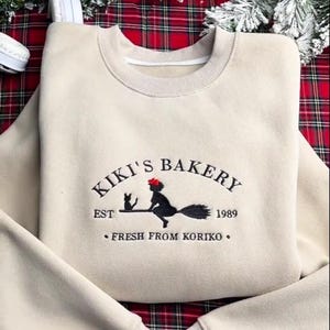 May include: A cream-coloured sweatshirt featuring the embroidered words "KIKI'S BAKERY" and a graphic of a witch on a broom with a black cat. The text "EST 1989" and "FRESH FROM KORIKO" are also embroidered.