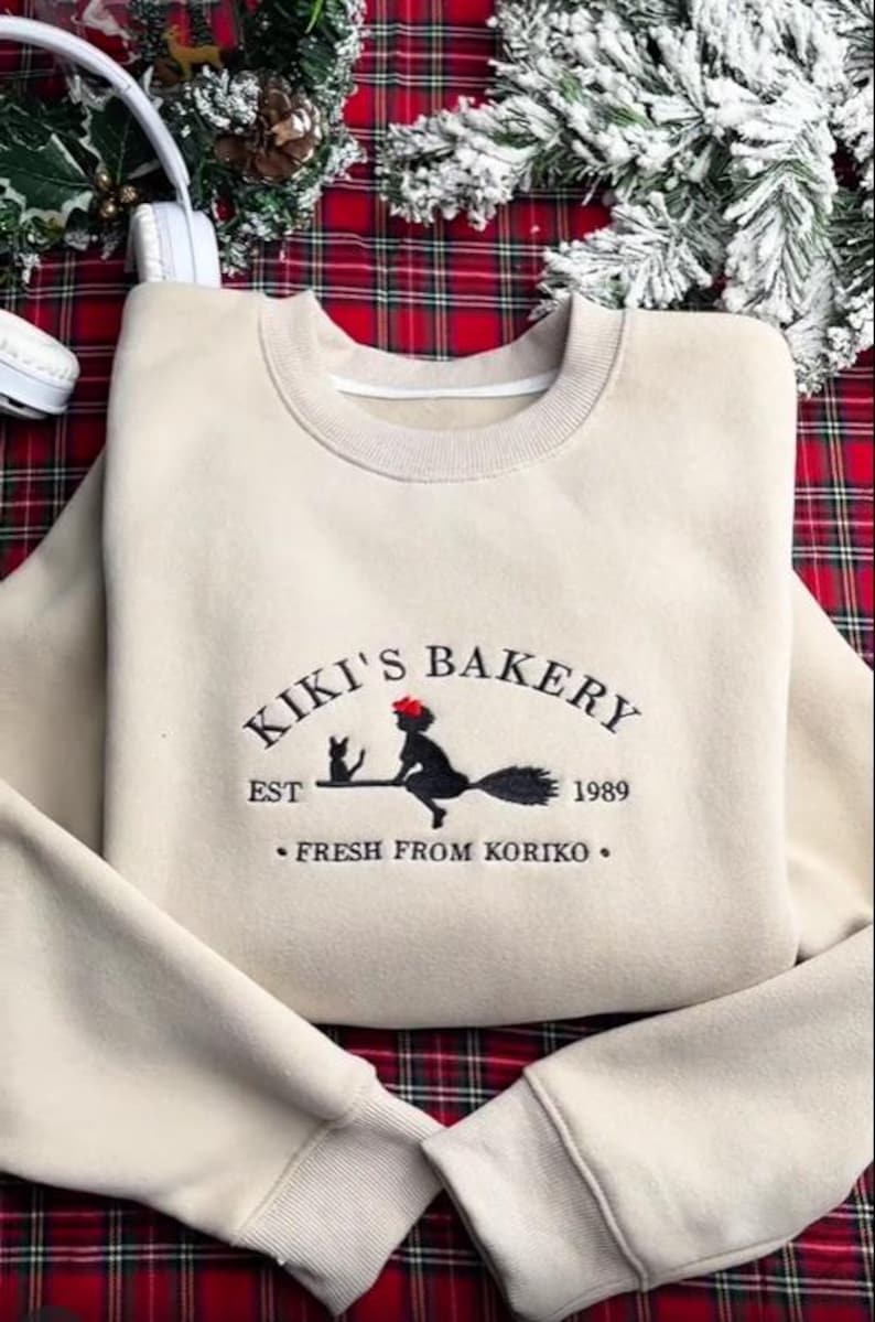 May include: A cream-coloured sweatshirt featuring the embroidered words "KIKI'S BAKERY" and a graphic of a witch on a broom with a black cat. The text "EST 1989" and "FRESH FROM KORIKO" are also embroidered.