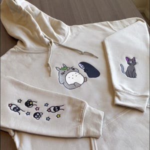 May include: Beige hoodie with embroidered cartoon characters. The front features Totoro and No-Face, while the sleeves display soot sprites and stars. A gray cat with a purple bow is on the right sleeve. The hoodie has a drawstring hood.