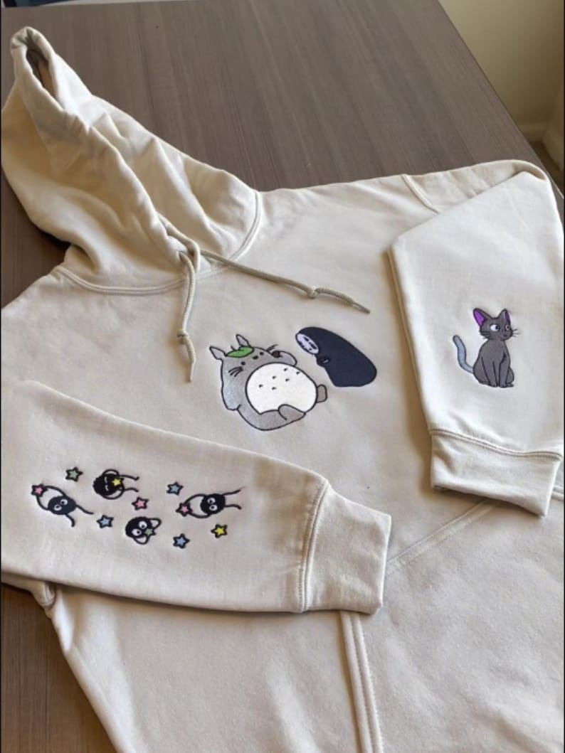 May include: Beige hoodie with embroidered cartoon characters. The front features Totoro and No-Face, while the sleeves display soot sprites and stars. A gray cat with a purple bow is on the right sleeve. The hoodie has a drawstring hood.