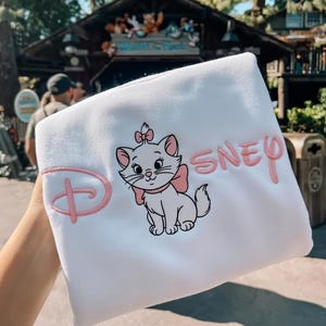 Peut inclure: Sweat-shirt blanc avec une image brod&eacute;e de Marie, le chat des Aristochats de Disney, avec un n&oelig;ud rose et le mot "Disney". Le logo Disney est &eacute;galement brod&eacute; en rose.