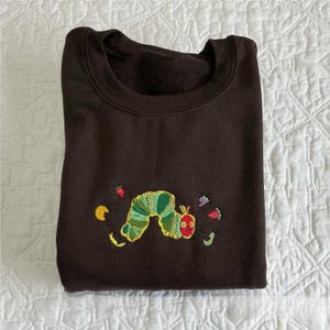 May include: A dark brown sweatshirt with an embroidered design of a green and yellow caterpillar with a red head. The caterpillar is surrounded by embroidered images of fruit and leaves.