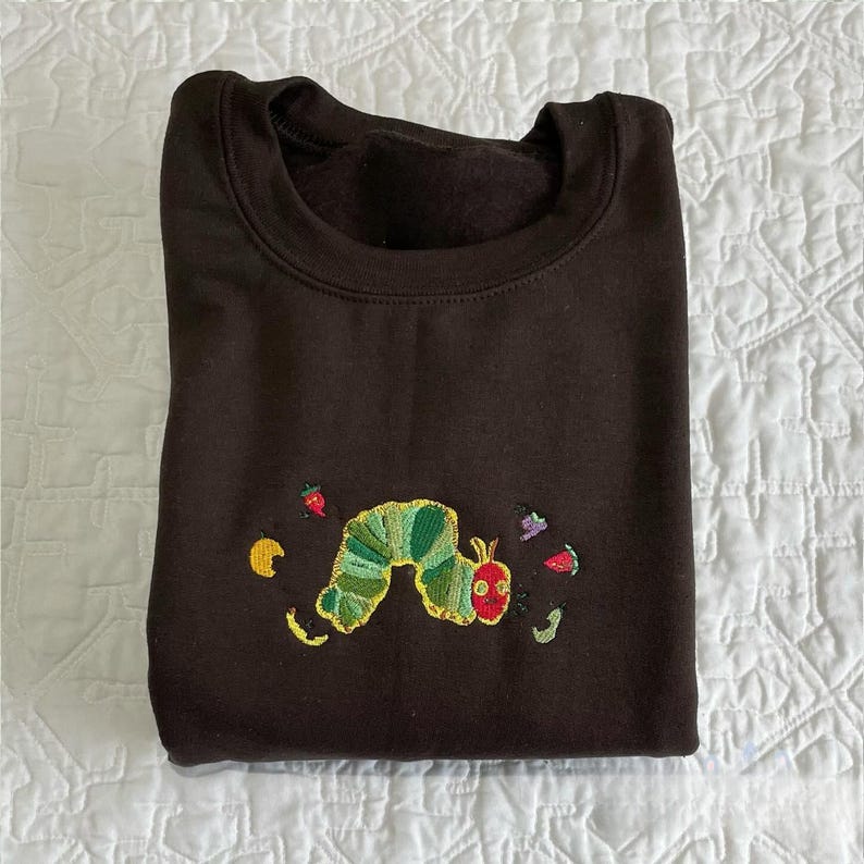 May include: A dark brown sweatshirt with an embroidered design of a green and yellow caterpillar with a red head. The caterpillar is surrounded by embroidered images of fruit and leaves.