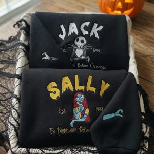May include: Two black sweatshirts featuring embroidered designs. The top sweatshirt displays white text reading JACK, a skeleton character, and the text Est. 1993. The bottom sweatshirt features yellow text reading SALLY, a female character, and Est. 1993.