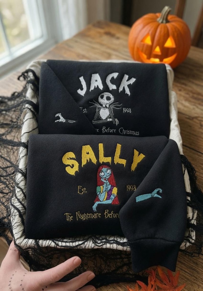 May include: Two black sweatshirts featuring embroidered designs. The top sweatshirt displays white text reading JACK, a skeleton character, and the text Est. 1993. The bottom sweatshirt features yellow text reading SALLY, a female character, and Est. 1993.