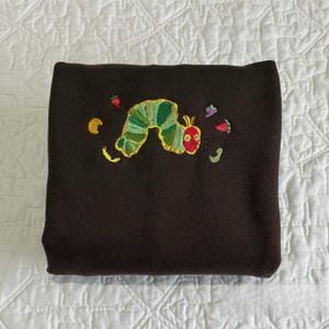 May include: A folded dark brown sweatshirt with an embroidered design of a green and yellow caterpillar with a red head. The caterpillar is surrounded by embroidered fruits and leaves in various colors.