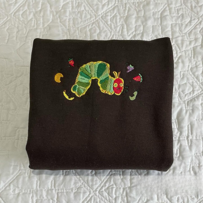 May include: A folded dark brown sweatshirt with an embroidered design of a green and yellow caterpillar with a red head. The caterpillar is surrounded by embroidered fruits and leaves in various colors.