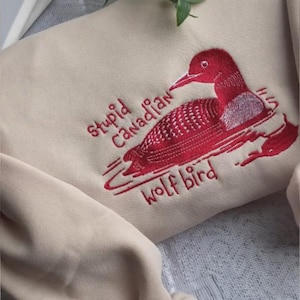 Pode incluir: Camisola bege com bordado vermelho de um mergulhão e o texto "Stupid Canadian Wolf Bird". A camisola está dobrada sobre uma superfície estampada.