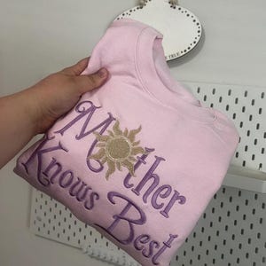 以下が含まれることがあります： 薄いピンク色のスウェットシャツ。紫色の筆記体で「Mother Knows Best」と刺繍されています。「ther」という単語の上には金の太陽のグラフィックがあります。白い壁を背景にスウェットシャツが展示されています。