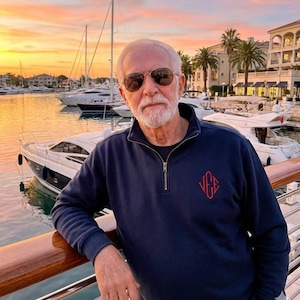 Könnte beinhalten: Ein Mann mit Sonnenbrille und einem marineblauen Pullover mit einem Viertelreißverschluss und einem roten Monogramm. Der Hintergrund zeigt einen Yachthafen mit Booten, Palmen und Gebäuden bei Sonnenuntergang.