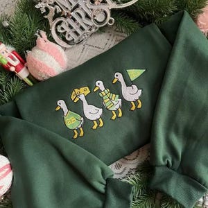 May include: A dark green sweatshirt with embroidered ducks, each wearing a different accessory. The ducks are in a row, with a Christmas tree and cheese wedge. The sweatshirt is surrounded by Christmas decorations.
