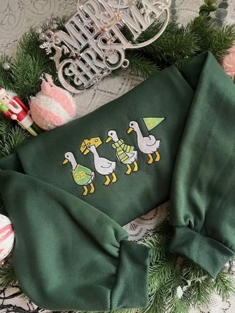 May include: A dark green sweatshirt with embroidered ducks, each wearing a different accessory. The ducks are in a row, with a Christmas tree and cheese wedge. The sweatshirt is surrounded by Christmas decorations.