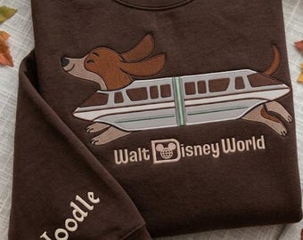 May include: A dark brown crewneck sweatshirt featuring an embroidered graphic of a dachshund dog inside a monorail train. The text Walt Disney World is embroidered below the graphic, and the name Noodle is embroidered on the sleeve cuff.