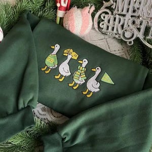 May include: A dark green sweatshirt featuring an embroidered design of four ducks in a row, each wearing a different outfit. The ducks are in yellow, white, and green. The sweatshirt is surrounded by Christmas decorations.