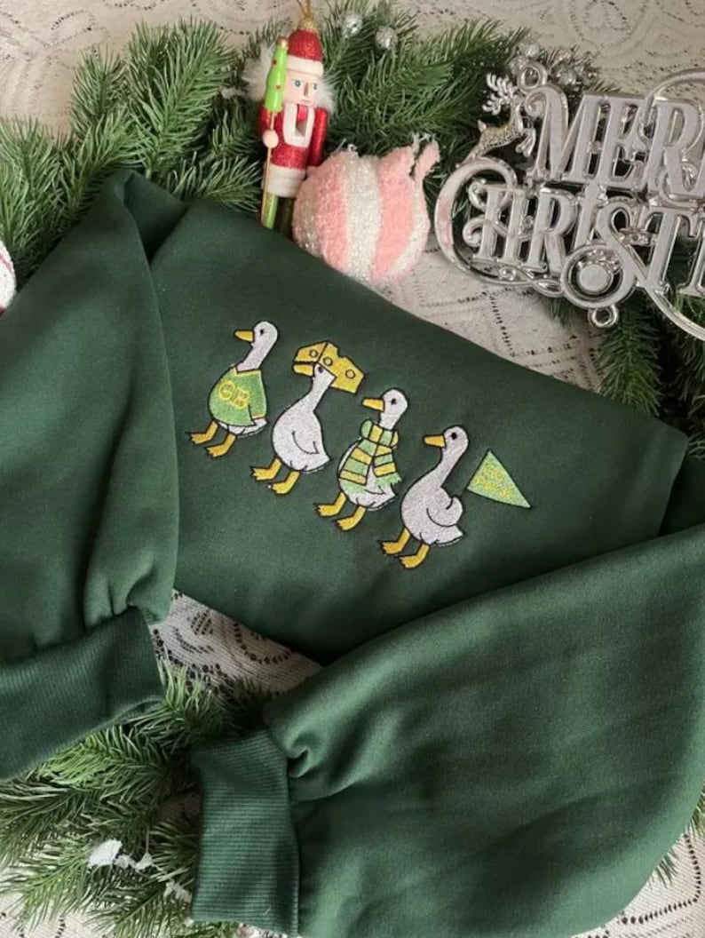 May include: A dark green sweatshirt featuring an embroidered design of four ducks in a row, each wearing a different outfit. The ducks are in yellow, white, and green. The sweatshirt is surrounded by Christmas decorations.
