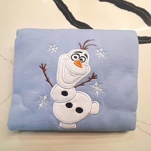 May include: A light blue blanket featuring an embroidered image of Olaf the snowman from the movie Frozen. The design includes white snowflakes and Olaf's face, body, and twig arms. The blanket is folded and appears soft.