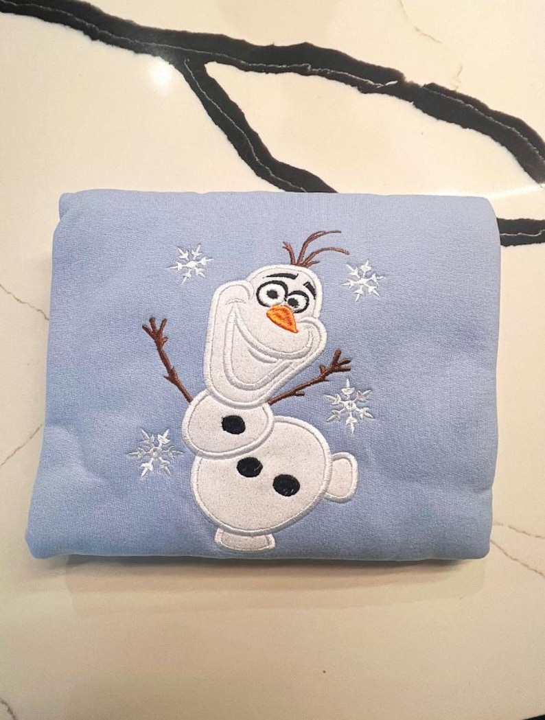 May include: A light blue blanket featuring an embroidered image of Olaf the snowman from the movie Frozen. The design includes white snowflakes and Olaf's face, body, and twig arms. The blanket is folded and appears soft.
