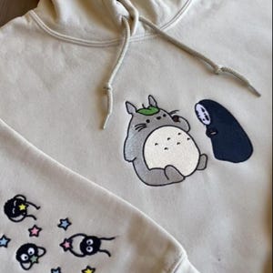 May include: Cream-colored hoodie featuring embroidered cartoon characters. The front displays a gray Totoro with a leaf and a black figure. The sleeve has additional cartoon figures and stars. The hoodie has a drawstring and ribbed cuffs.