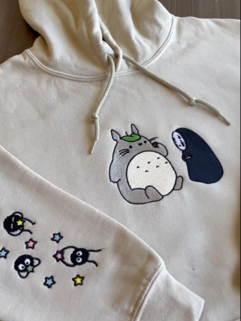 May include: Cream-colored hoodie featuring embroidered cartoon characters. The front displays a gray Totoro with a leaf and a black figure. The sleeve has additional cartoon figures and stars. The hoodie has a drawstring and ribbed cuffs.