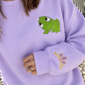 Pode incluir: Uma camisola de gola redonda em violeta claro com um camaleão verde bordado no peito e um pequeno design de coroa colorido com detalhes em violeta e amarelo bordado no punho da manga.