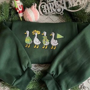 May include: Dark green sweatshirt with embroidered ducks wearing accessories. The ducks are wearing a cheese hat, a scarf, and a flag. The sweatshirt has long sleeves and a crew neck. Christmas decorations are in the background.