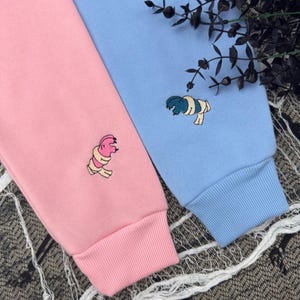 May include: A light pink long-sleeve sweatshirt and a light blue long-sleeve sweatshirt, each featuring a small embroidered graphic of a mummy character. The pink sweatshirt has a pink mummy, and the blue sweatshirt has a dark blue mummy.