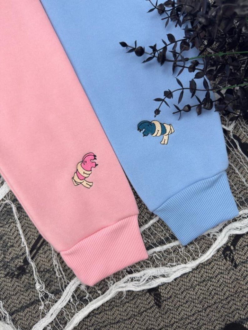May include: A light pink long-sleeve sweatshirt and a light blue long-sleeve sweatshirt, each featuring a small embroidered graphic of a mummy character. The pink sweatshirt has a pink mummy, and the blue sweatshirt has a dark blue mummy.