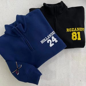 May include: Two quarter-zip pullovers, one navy blue and one black. The navy blue pullover has "HOLLANDER 24" embroidered in white, and hockey sticks on the sleeve. The black pullover has "ROZANOV 81" embroidered in yellow.