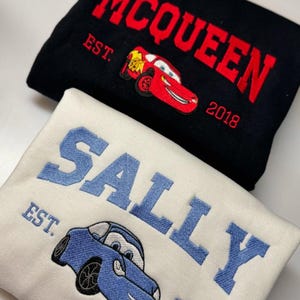 May include: Two sweatshirts, one black and one cream, with car embroidery. The black sweatshirt has "MCQUEEN" in red, a red car graphic, and "EST. 2018." The cream sweatshirt features "SALLY" in blue, a blue car graphic, and "EST. 2018."