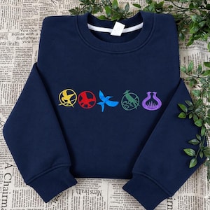 May include: A navy blue crewneck sweatshirt featuring five embroidered circular icons across the chest. The designs include a yellow mockingjay, a red bird, a blue bird, a green bird, and a purple floral-like shape, all centred on the fabric.