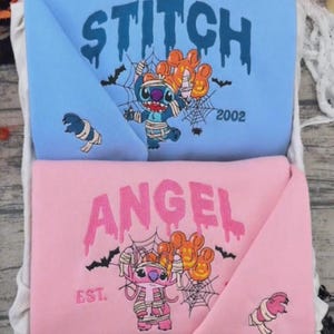 May include: A light blue sweatshirt featuring a blue Stitch graphic wrapped in bandages with orange balloons and the text STITCH 2002, and a pink sweatshirt featuring a pink Angel graphic wrapped in bandages with orange balloons and the text ANGEL EST.