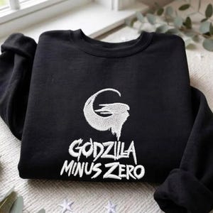 Puede incluir: Una sudadera negra de manga larga con bordado blanco en el pecho. El diseño incluye un logotipo de letra G estilizada sobre el texto "GODZILLA MINUS ZERO" en una fuente dentada y desgastada. La prenda tiene cuello redondo y puños acanalados.
