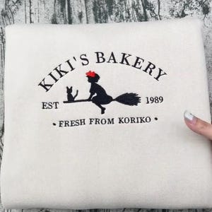 May include: Cream-coloured sweatshirt featuring a black graphic of a witch on a broom with a cat, and the text "Kiki's Bakery" and "Fresh from Koriko". The year "1989" is also displayed.