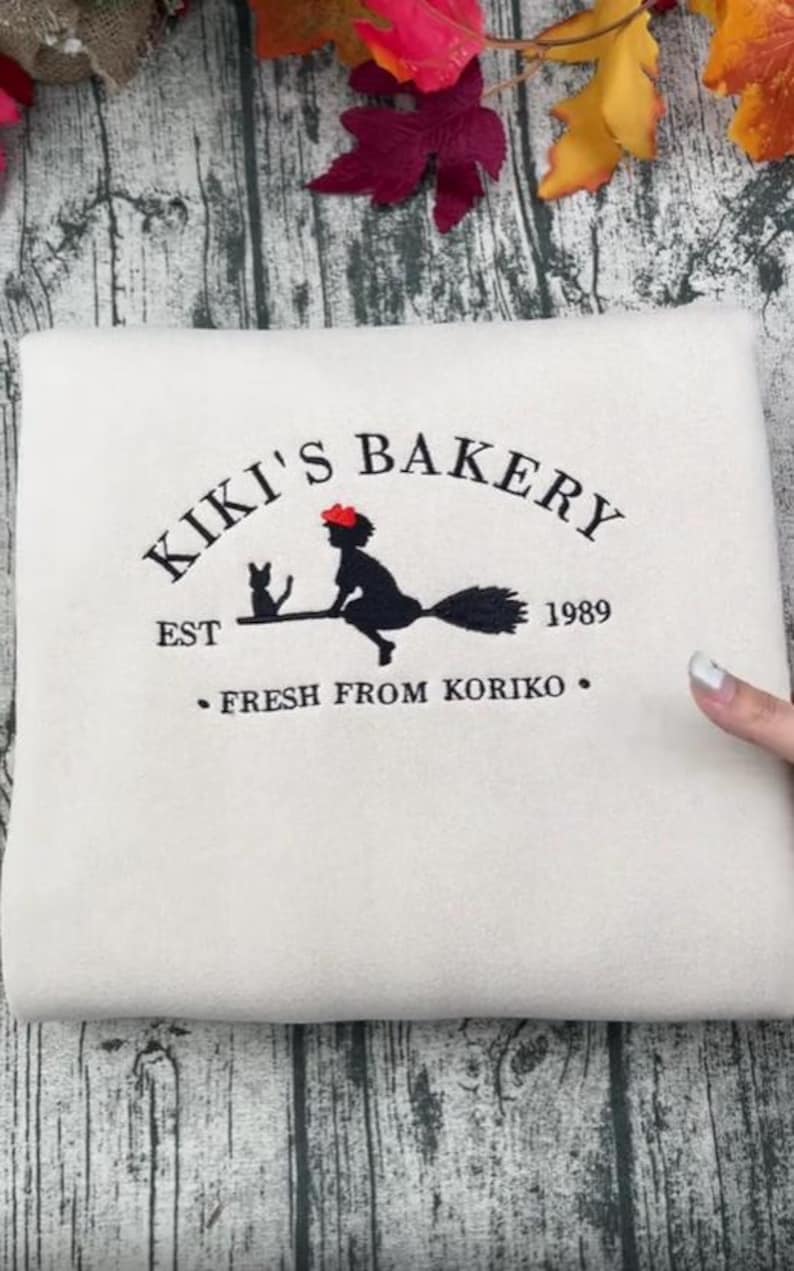 May include: Cream-coloured sweatshirt featuring a black graphic of a witch on a broom with a cat, and the text "Kiki's Bakery" and "Fresh from Koriko". The year "1989" is also displayed.