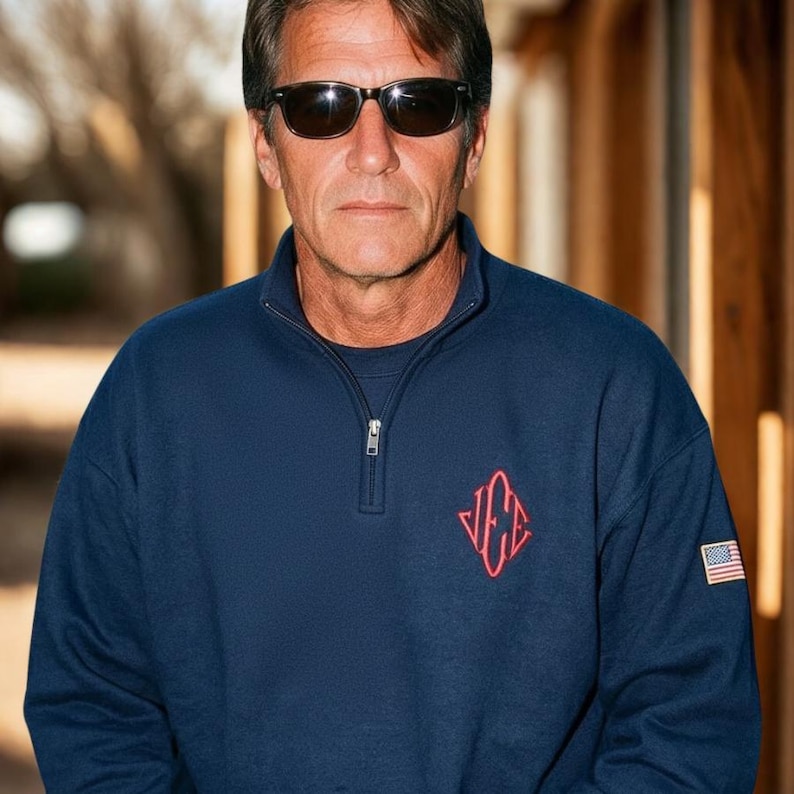 May include: Navy blue sweatshirt with a quarter-zip collar and a red emblem on the chest. The sweatshirt features a small American flag patch on the sleeve. The person in the image is wearing black sunglasses.
