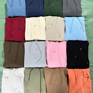 Peut inclure: Une collection de sweats &agrave; capuche pli&eacute;s de diff&eacute;rentes couleurs, dont le bleu, le vert, le rouge et le rose. Chaque sweat &agrave; capuche a une capuche &agrave; cordon de serrage et un petit d&eacute;tail circulaire rouge pr&egrave;s de la poitrine. Les sweats sont dispos&eacute;s avec soin.