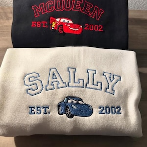 Może przedstawiać: Dwie złożone bluzy, jedna czarna i jedna biała. Czarna bluza ma czerwony haft "MCQUEEN" z czerwoną grafiką samochodu i "EST. 2002". Biała bluza ma niebieski haft "SALLY" z niebieską grafiką samochodu i "EST. 2002".