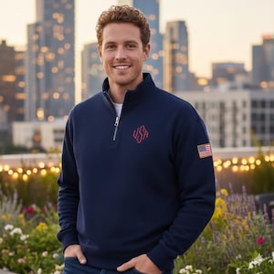 May include: Navy blue quarter-zip sweatshirt with a red "USA" logo and an American flag patch on the sleeve. The sweatshirt has a white undershirt and is made of a soft material. The model is wearing blue jeans.