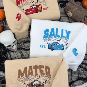 May include: Three sweatshirts in beige and white featuring embroidered designs of characters from the movie Cars. Each sweatshirt displays a character name, a car illustration, spider web details, and a small heart-shaped patch on the sleeve.
