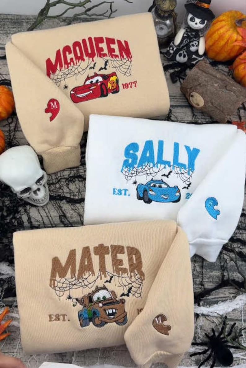 May include: Three sweatshirts in beige and white featuring embroidered designs of characters from the movie Cars. Each sweatshirt displays a character name, a car illustration, spider web details, and a small heart-shaped patch on the sleeve.