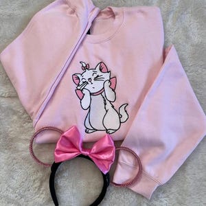 K&ouml;nnte beinhalten: Hellrosa Sweatshirt mit gestickter wei&szlig;er Katze und rosa Akzenten. Ein passendes rosa Schleifen-Stirnband mit schwarzem Band und glitzernden Ohren liegt neben dem Sweatshirt. Die Katze ist aus Aristocats.